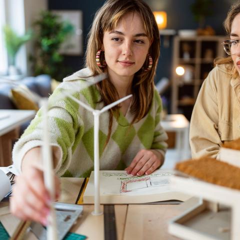 Two young women work at a desk, looking at a scale model of a wind turbine