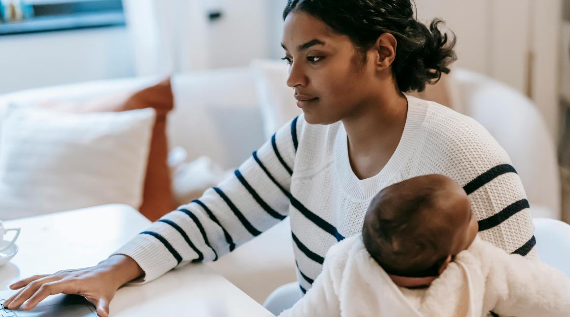 A woman holds her baby while working from home on a laptop