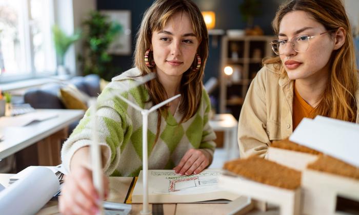 Two young women work at a desk, looking at a scale model of a wind turbine