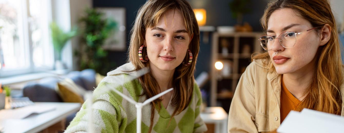 Two young women work at a desk, looking at a scale model of a wind turbine